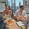 Woman and Man are laughing while working at their desk together while eating lunch with a view of the city behind them 
